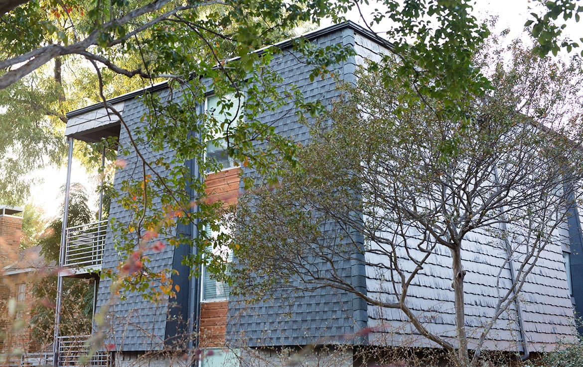 A modern building with shingled siding and large windows is surrounded by leafy trees. The sunlight filters through the branches, casting dappled shadows on the building. The structure features a balcony with metal railings on the left side.