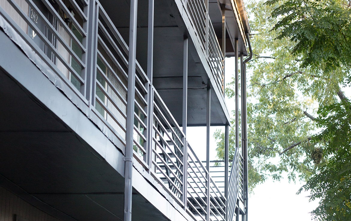 A perspective view of the exterior of a multi-story building with metal railings and walkways on each floor. Tall trees with lush green leaves are visible to the right of the building, under a clear sky.