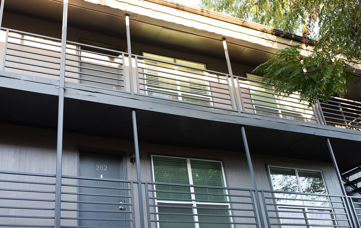 A two-story apartment building with grey walls and matching metal railings. The door of unit 202 is visible on the lower level, next to two windows with closed blinds. A tree with green leaves partially covers the view on the right.