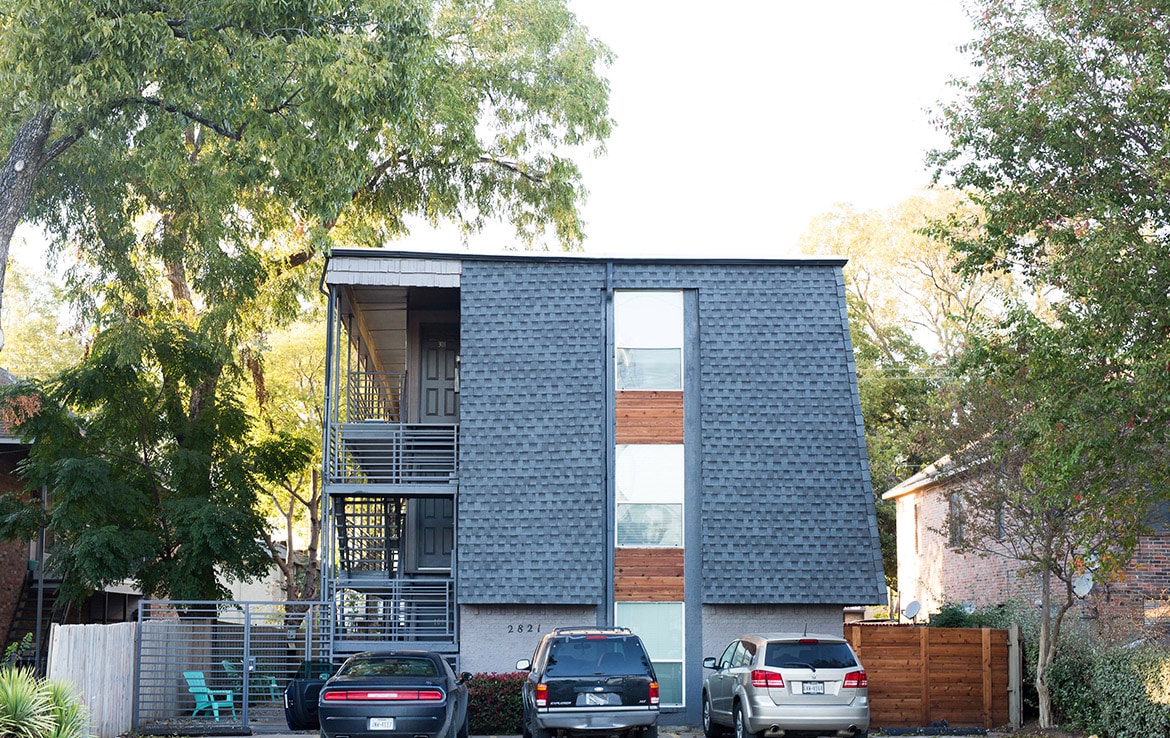 A modern apartment building with a unique architectural design featuring gray textured exterior walls and wooden accents. Three floors are visible with balconies on the left side. Two cars and an SUV are parked in front. Tall trees partially shade the building.