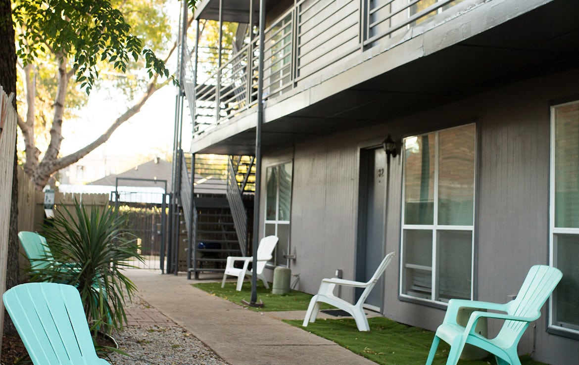 A quiet apartment complex with a two-story building, metal railings, and an exterior staircase. There are white and turquoise plastic chairs placed along the walkway and patches of artificial grass outside the units. Trees provide shade from the sun.