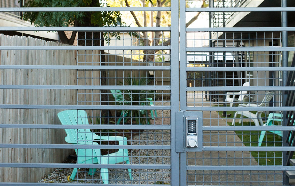 A gated outdoor area features light blue and white Adirondack chairs on gravel and grass surfaces. A keypad lock is on the gate in the foreground. Wooden fencing and a building with a walkway are visible in the background.