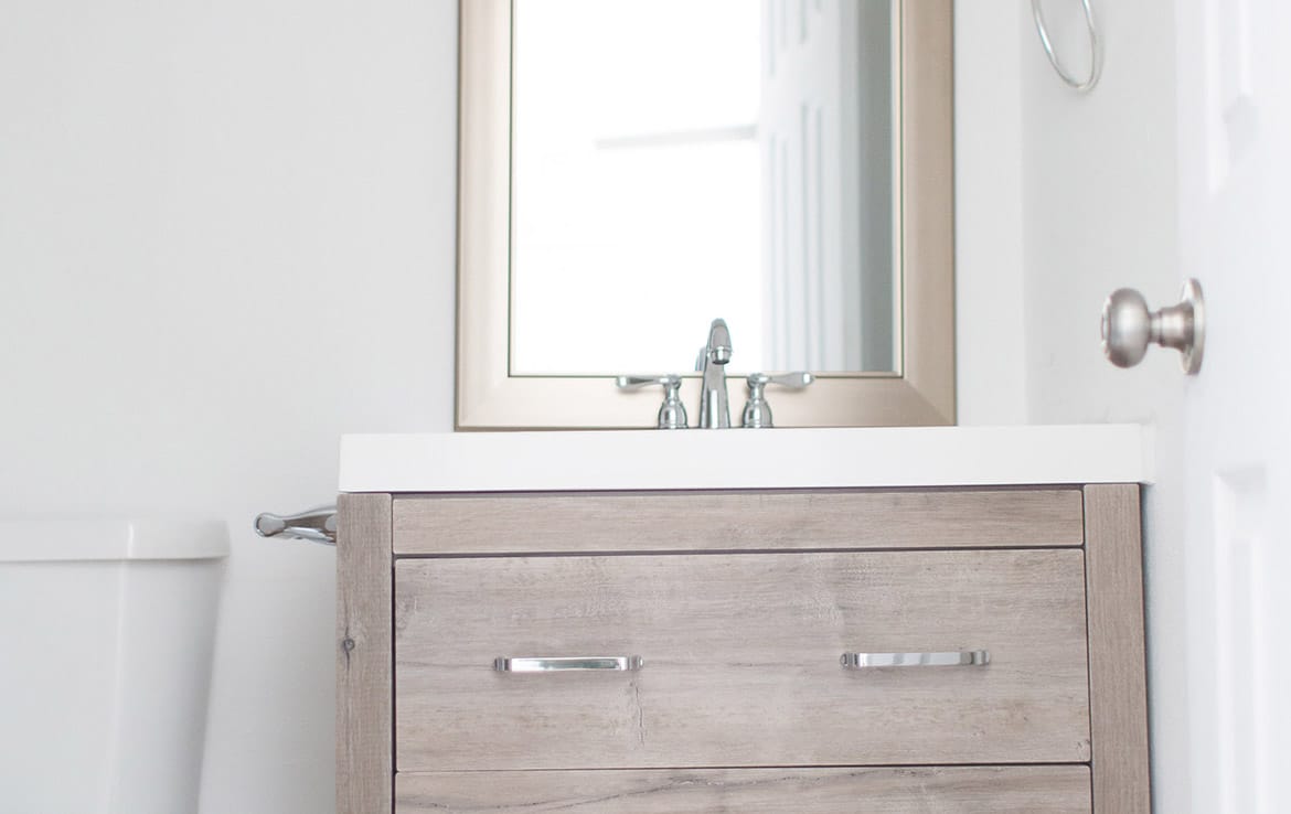 A minimalist bathroom featuring a wooden vanity with chrome handles, a white countertop with a silver faucet, and a rectangular mirror with a light-colored frame. There is a towel ring on the wall and a glimpse of a toilet to the left.