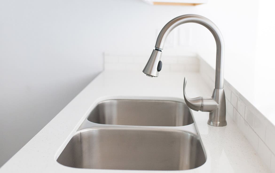 A modern kitchen sink with a dual-basin stainless steel design and a sleek, curved stainless steel faucet on a white countertop. The background is a light-colored wall and a partial view of white subway tiles.