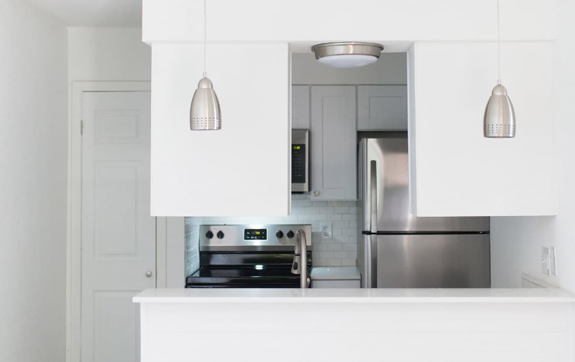 A modern kitchen with white cabinets, a stainless steel refrigerator, and an electric stove with a microwave above. Two pendant lights hang from the ceiling. The decor is minimalistic and the space is well-lit with a clean, sleek design.