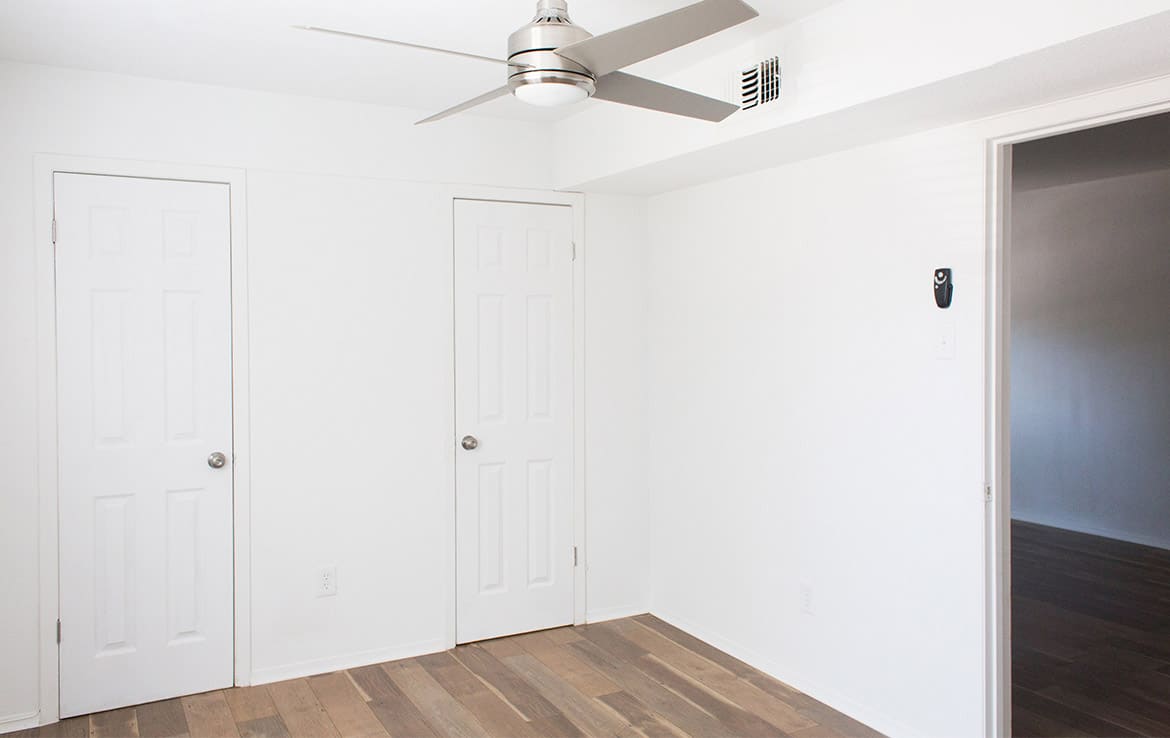 A minimalistic room with white walls features two white doors with silver knobs. A modern ceiling fan with a light is mounted on the ceiling. The floor is wooden, and there is an open doorway on the right leading to another room.