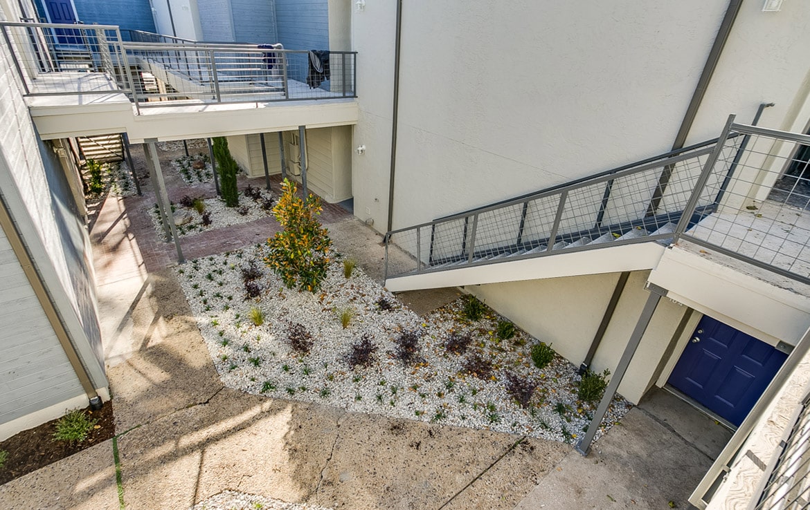 A view of a modern apartment complex from Our Portfolio at Orand Capital features gray railings and various staircases. The landscaped courtyard area boasts white gravel, small plants, and shrubs. The light-colored buildings include a door painted in blue, adding a touch of color to the serene environment.