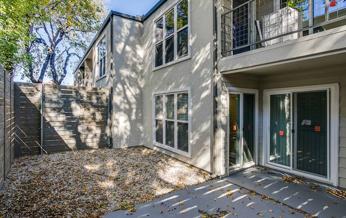 A two-story building with a fenced patio area covered in fallen leaves is one of the highlights of our portfolio at Orand Capital. The building features multiple large windows and sliding glass doors. Trees surround the area, casting shadows on the exterior wall and patio.