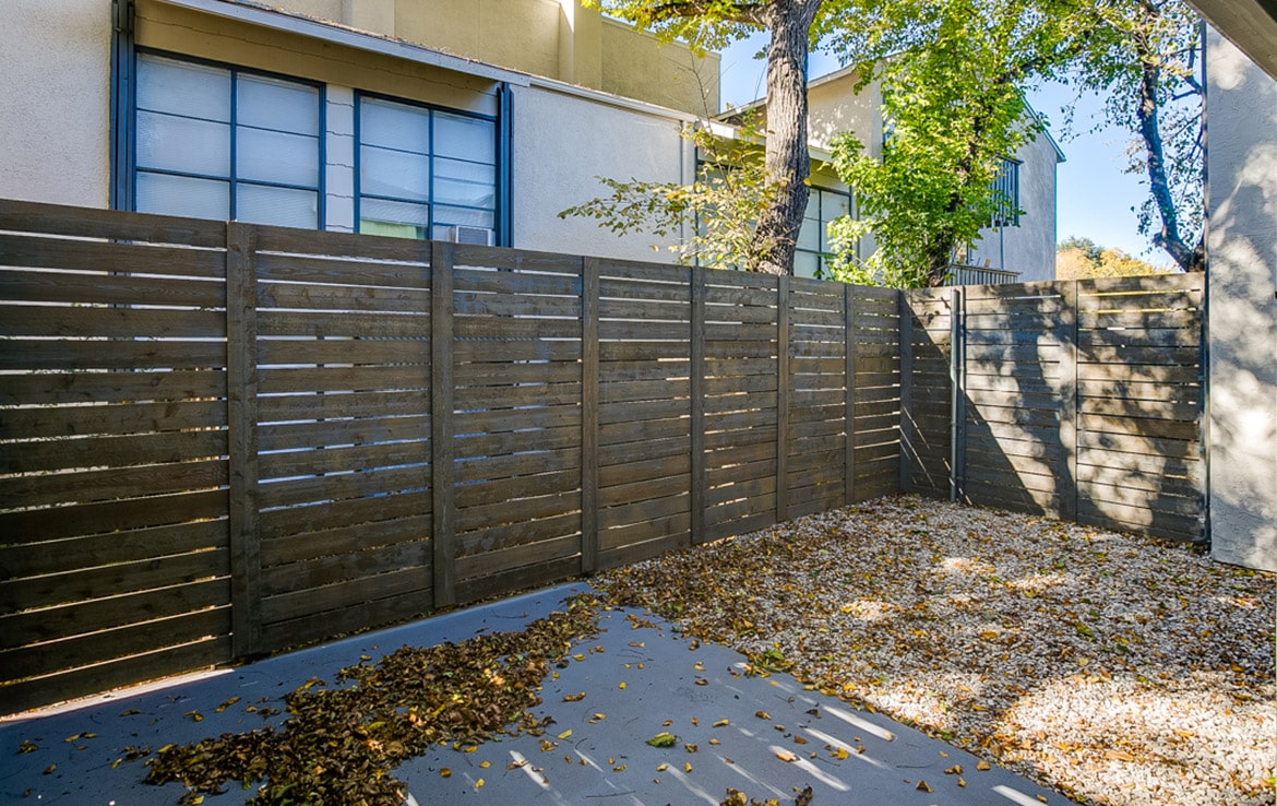 A backyard with a wooden slat fence enclosing a small area. The ground is a mix of gravel and a concrete patio, with fallen leaves scattered around. Trees with green and yellow leaves stand near the fence, while buildings rise in the background—an inviting snapshot from our portfolio at Orand Capital.