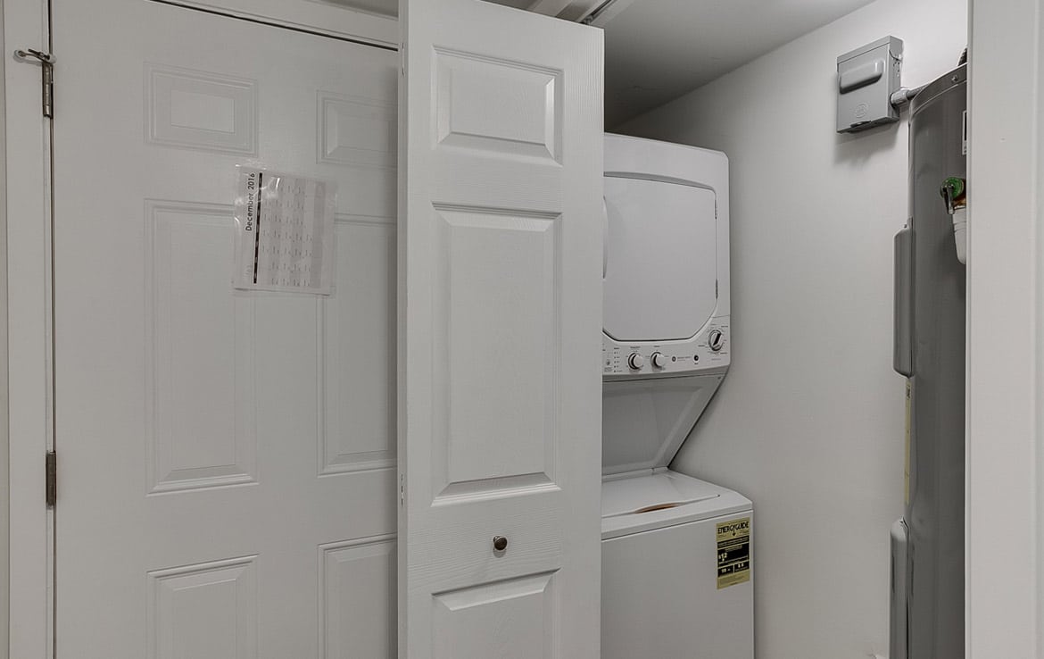 A small laundry nook with white folding doors partially open, revealing a stacked washer and dryer unit. The space is compact and located next to a water heater. The walls and doors are painted white, creating a clean and bright appearance, exemplifying the efficient designs in Our Portfolio - Orand Capital.
