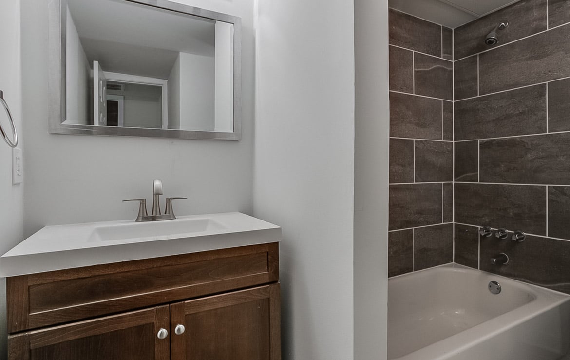 A modern bathroom with a rectangular mirror above a white sink and a wooden vanity. To the right is a bathtub with grey tile walls and silver fixtures. The bathroom has a clean, minimalist design and is featured in our portfolio at Orand Capital.