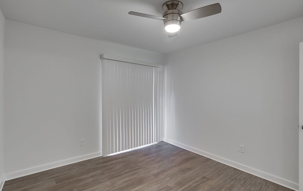 A small, empty room with light gray walls and wood flooring. The room features a ceiling fan with a light, and vertical blinds covering a sliding glass door that allows natural light to enter. The clean, minimalistic design is something you'd find in Our Portfolio at Orand Capital.