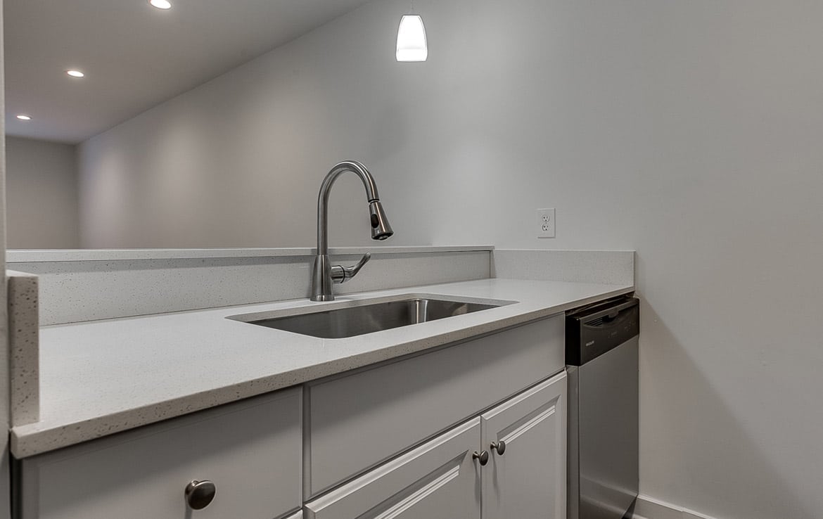 Modern kitchen with a sleek design featuring a white countertop and cabinetry. A stainless steel sink with a high-arc faucet is centered in the counter. A stainless steel dishwasher is built into the cabinetry on the right side. Soft overhead lighting enhances this elegant space within Our Portfolio - Orand Capital.
