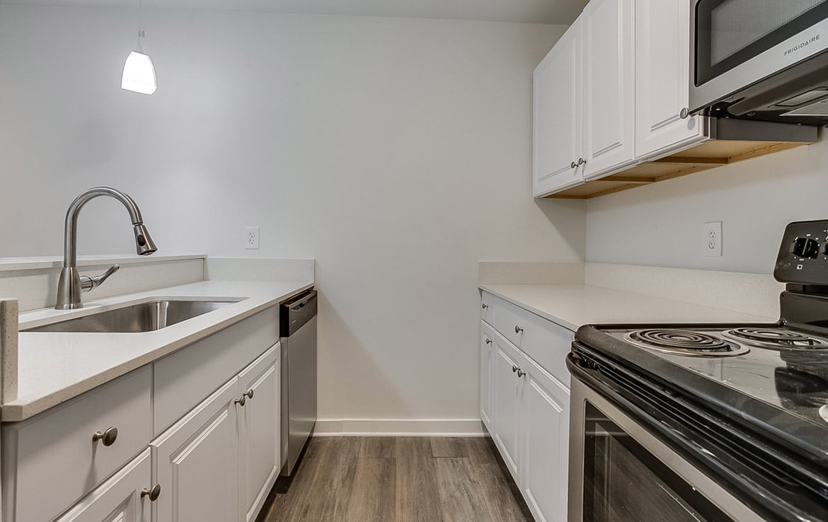 A modern kitchen from our portfolio, Orand Capital, featuring white cabinets, a stainless steel sink with a pull-down faucet, a built-in dishwasher, and an electric stove with a black top. The light-colored countertops complement the wood flooring. A pendant light elegantly hangs above the sink.