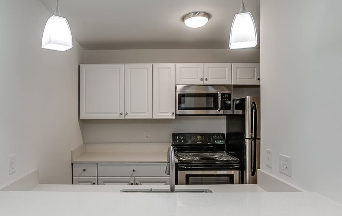 A modern kitchen with white cabinets, stainless steel appliances including a microwave and oven, and a double-door refrigerator. The countertop is white with a minimalist design, illuminated by two pendant lights hanging from the ceiling. This remarkable space is featured in Our Portfolio - Orand Capital.