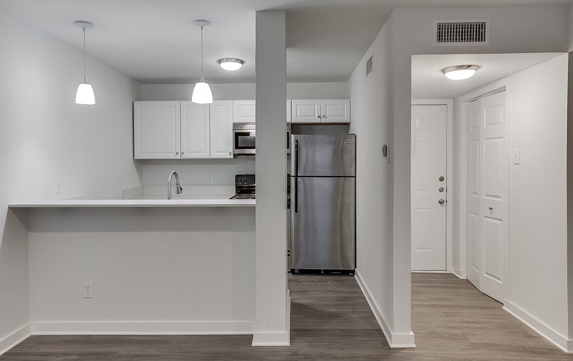 A modern, minimalist kitchen from our portfolio at Orand Capital features white cabinetry, a stainless steel refrigerator, and a built-in microwave. The kitchen island boasts a white countertop and two hanging pendant lights. Wood flooring enhances the bright, clean appearance, with an entryway visible on the right.
