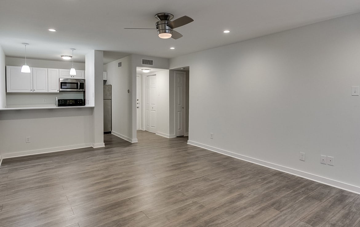 A modern, unfurnished apartment interior with an open layout, featuring a ceiling fan above a spacious living area with grey wood floors. The background shows a white kitchen with stainless steel appliances, including a refrigerator, microwave, and oven. This stylish space is part of Our Portfolio at Orand Capital.