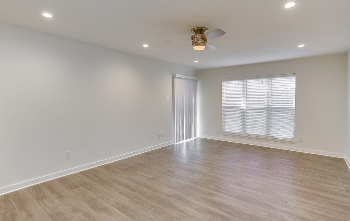 A bright, empty room with white walls and wooden flooring. A modern ceiling fan with a light is centered on the ceiling. A window with closed blinds is on the right and a sliding glass door with vertical blinds is located on the left side of the room, showcasing our portfolio's versatility at Orand Capital.