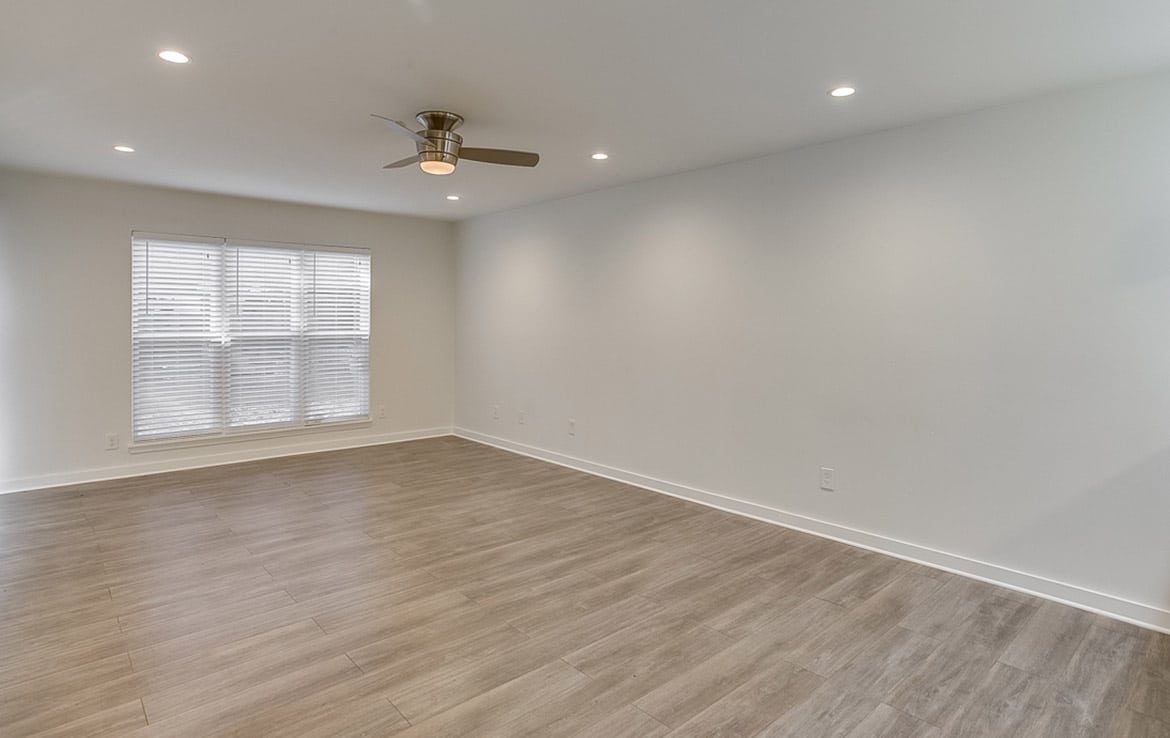 A modern, empty room with light gray hardwood flooring, white walls, and recessed ceiling lights. A large window with white blinds allows natural light to flood the space. A ceiling fan with a light fixture is centered on the ceiling. This elegant design is a standout in our portfolio at Orand Capital.