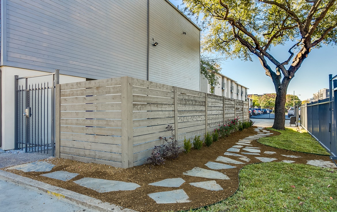 An outdoor area with a modern wooden fence, a stone pathway, and landscaped greenery. The pathway is lined with plants and the scene includes a nearby tree and a building with light gray siding. A metal gate is visible on the left side. Featured in Our Portfolio - Orand Capital.
