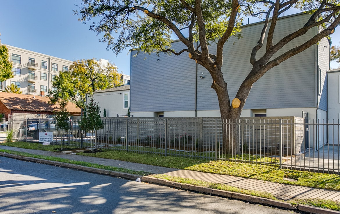 A light grey, two-story modern house with large windows is surrounded by a metal fence and trees. The property, featured in our portfolio - Orand Capital, is well-maintained with a green lawn and a pathway leading to the entrance. The background shows part of a residential neighborhood with other buildings.