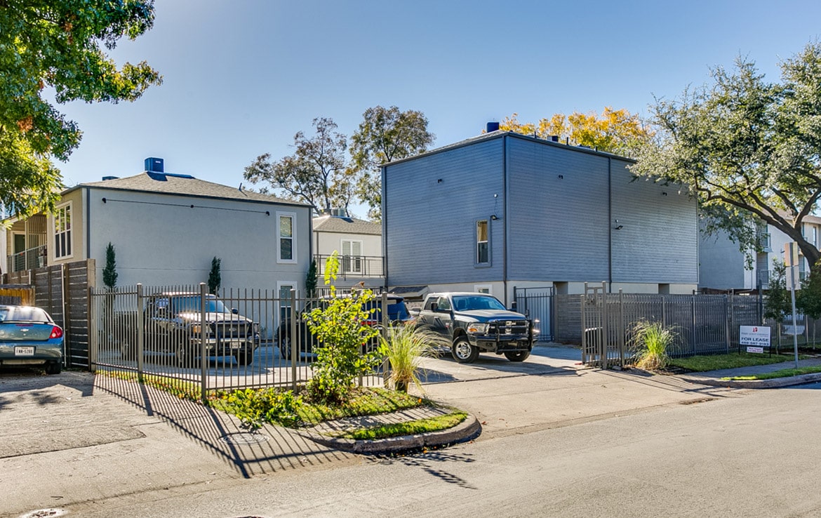 A suburban street features two modern houses behind a black metal fence, showcased in Our Portfolio - Orand Capital. The left house is light-colored with a pitched roof, while the right house is darker with a flat roof. Three vehicles sit in the driveways, surrounded by trees.