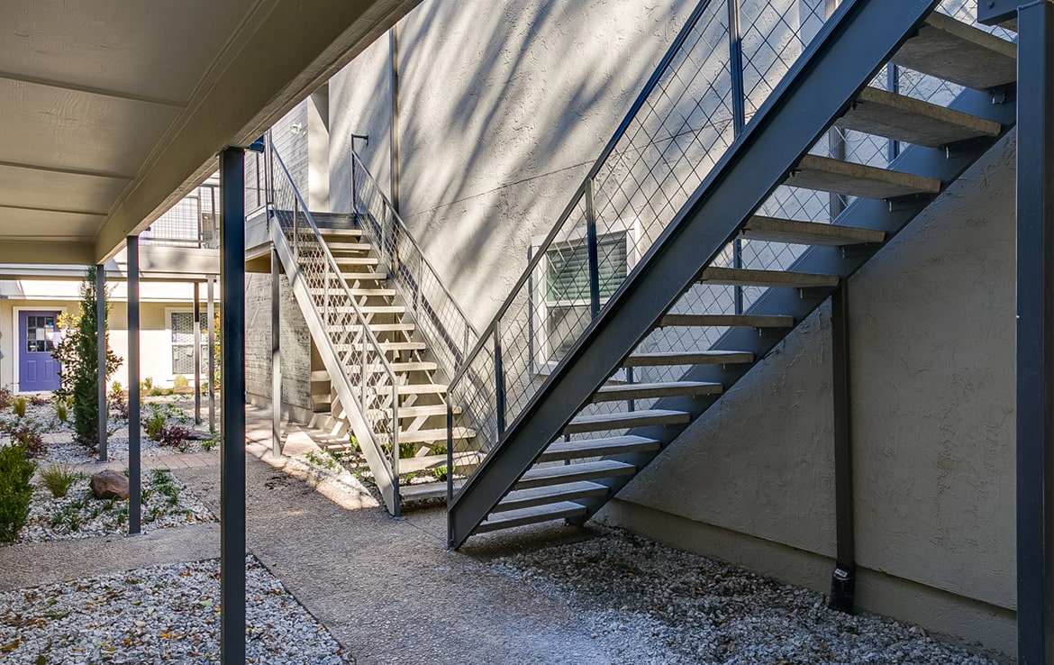 Exterior view of an apartment complex from Our Portfolio at Orand Capital. It features two sets of metal stairs leading to the upper floors - one to the left and one to the right. The area below is landscaped with gravel and patches of greenery, and the light-colored walls add a bright touch.