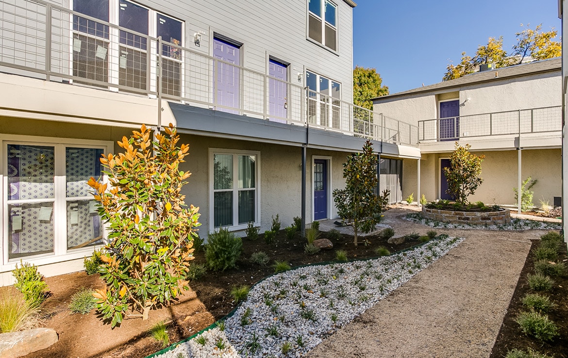 A modern apartment building with a two-story layout, featuring balconies and large windows. The courtyard has a landscaped garden with neatly paved pathways, various plants, and small trees. The exterior is painted gray with purple doors—another exquisite addition to our portfolio by Orand Capital.