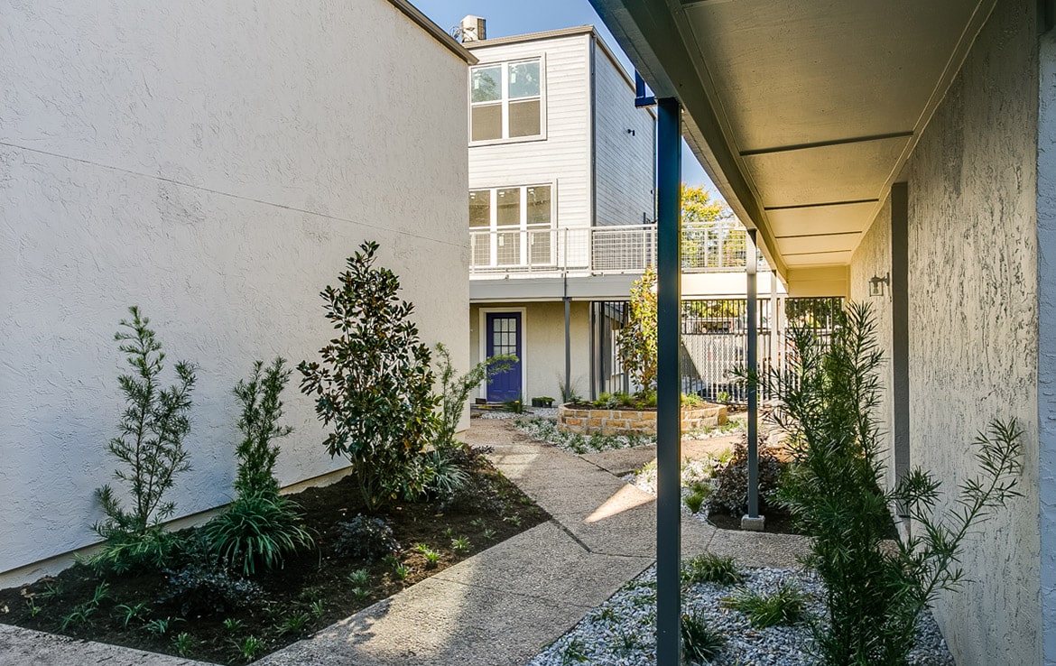 A narrow pathway lined with various plants and shrubs leads to a blue door of a modern two-story building with large windows and a balcony. The walls are painted white, and the garden area beside the path features gravel and small green plants. This charming entrance is part of Our Portfolio at Orand Capital.