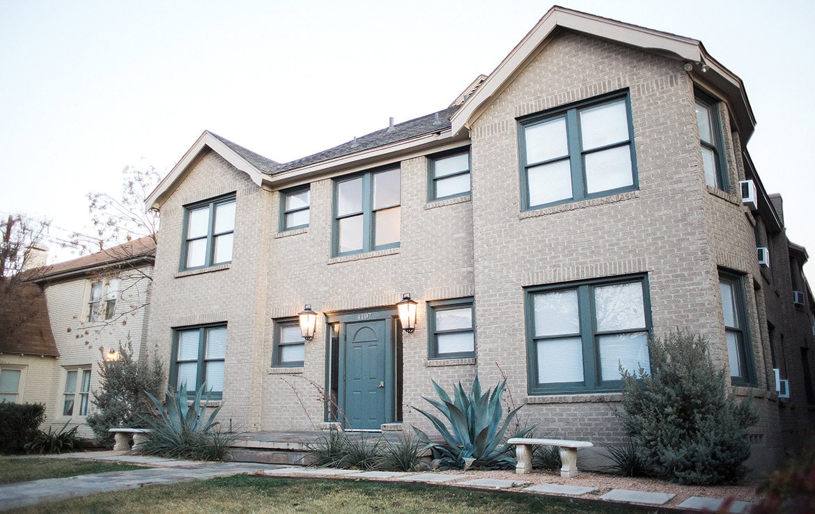 A two-story beige brick house with a teal door and teal window frames. The front yard has two large agave plants, shrubs, and concrete benches flanking the entryway. Two lamps frame the front door, illuminating the entrance—an exquisite addition to Our Portfolio at Orand Capital.