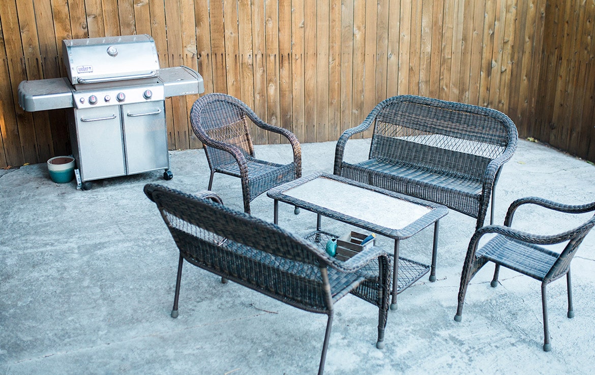 Discover our portfolio featuring a backyard patio with a wicker furniture set, including a loveseat, two chairs, and a coffee table arranged on a concrete floor. A stainless steel gas grill is positioned against a wooden fence in the background—an exquisite example from Orand Capital.