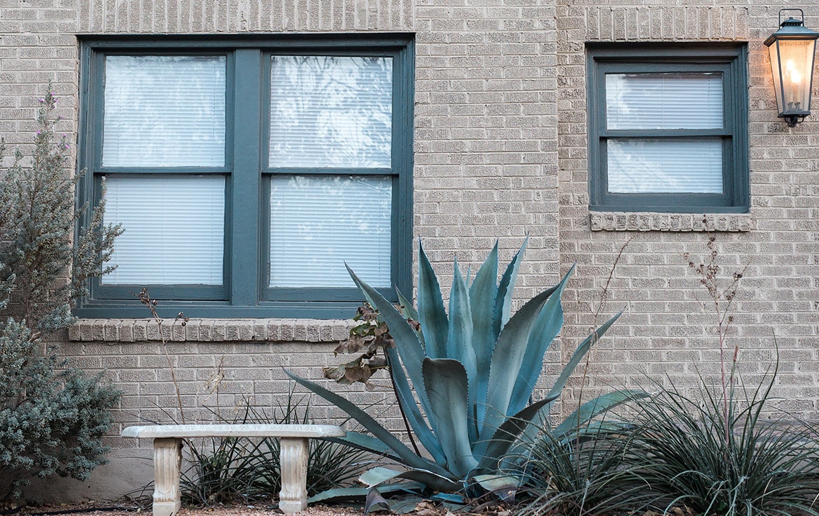A brick house with two windows, the left larger than the right. Each window has greenish-blue trim and white blinds. An outdoor lantern is mounted on the upper right. Below the left window is a stone bench; nearby is an agave plant surrounded by tall grasses—a charming addition to Our Portfolio - Orand Capital.