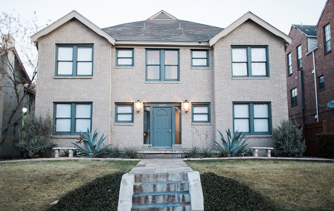 A two-story brick house with blue trim and a central entrance door flanked by two outdoor lights. There are steps leading up to the front door, with agave plants on either side of the walkway and well-maintained grass on the lawn, exemplifying a prime property in our portfolio at Orand Capital.