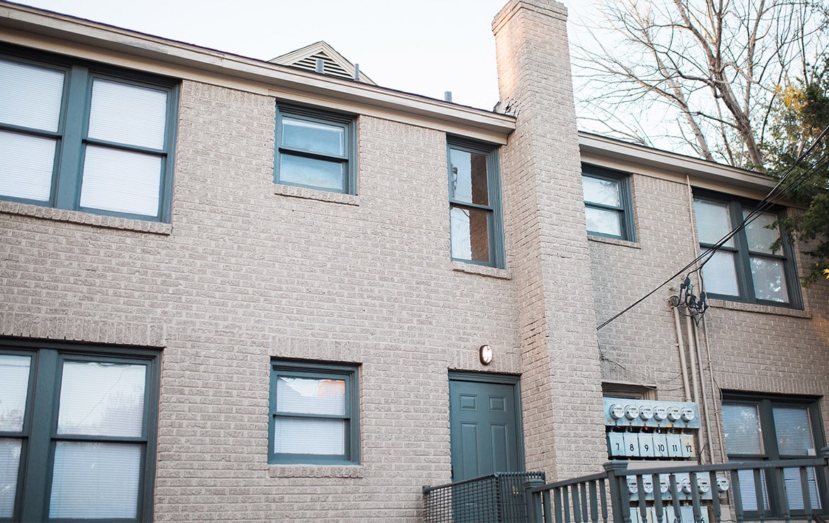 A brick apartment building with multiple windows and a green door stands tall. Near the entrance, multiple utility meters are mounted on the side. Some trees add a touch of nature in the background. The sky is clear, and the overall scene is well-lit. This charming property is part of Our Portfolio at Orand Capital.