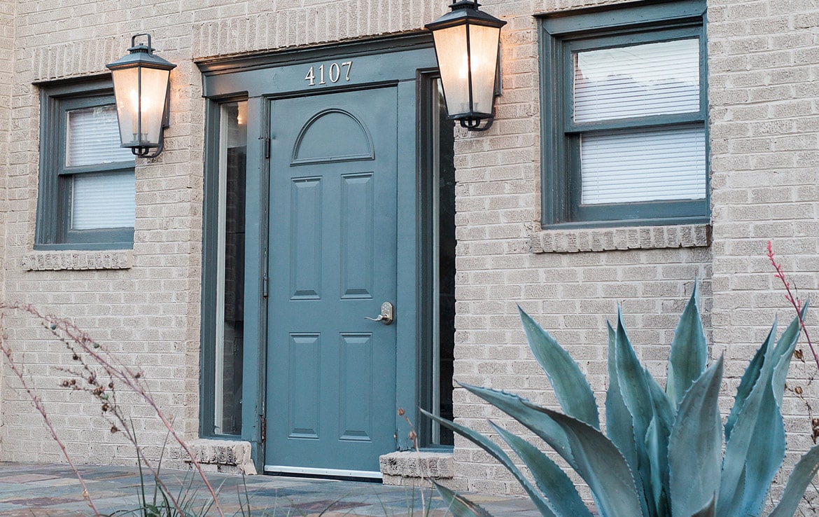 Our Portfolio: A beige brick building facade featuring a blue door with the number 4107 above it. The door is flanked by two rectangular windows with closed blinds and two black lantern-style lights. In front, there is a large agave plant on the right side—an exemplary property in Orand Capital's collection.