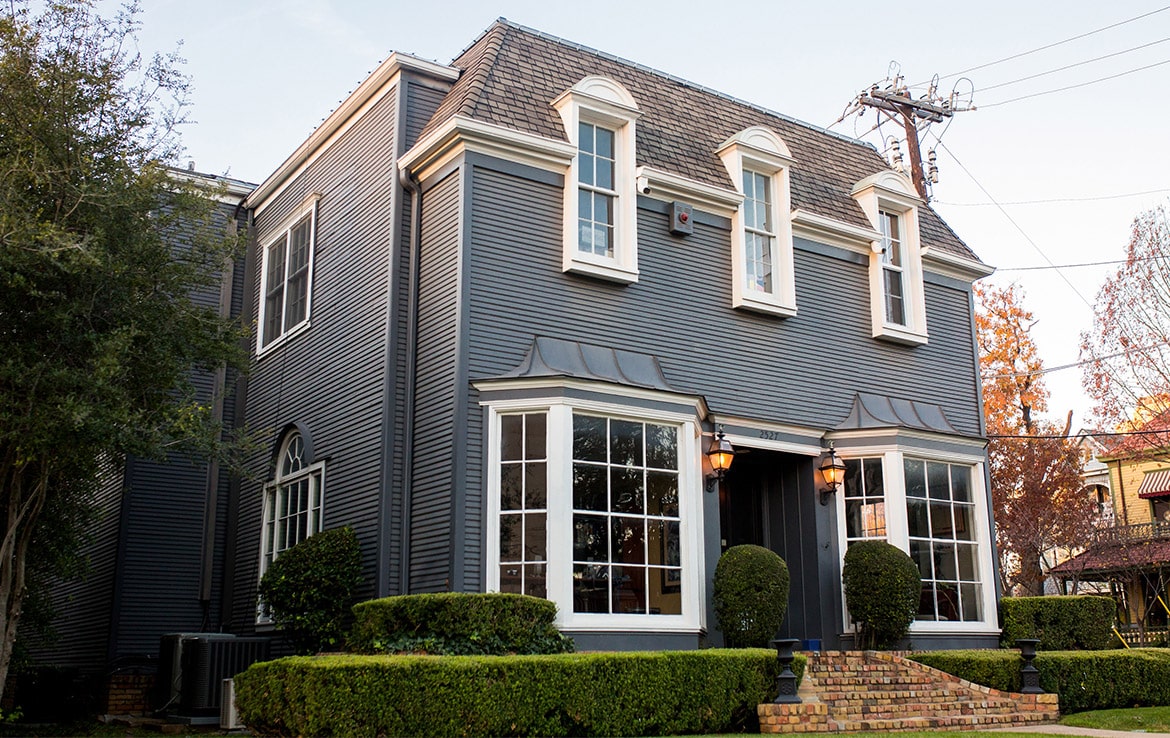 A two-story suburban house with dark grey siding, white trim, and a shingled roof. The facade features large bay windows, a black front door with lantern-style lights on either side, and well-kept green hedges in the front yard—another gem in Our Portfolio at Orand Capital.