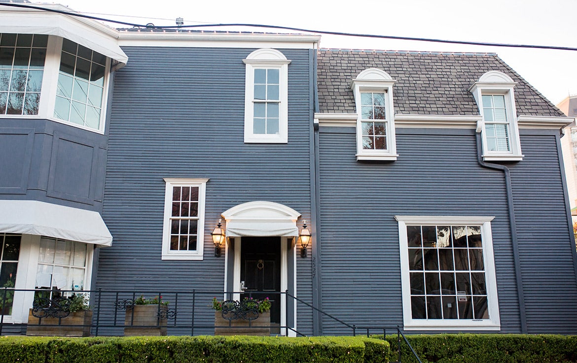A two-story house with gray siding, white trim, and a shingled roof. The front features two lantern-style lights flanking the door, several windows of varying sizes, and a small front hedged garden. The windows have flower boxes, and a power line runs across the image—part of our portfolio at Orand Capital.