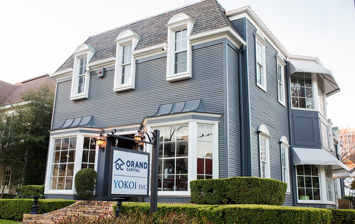 A large, two-story building painted in gray-blue with white trim features French-style windows and dormers on the roof. A sign outside reads "ORAND Capital" and "YOKOI Inc." The landscaped surrounding area includes trimmed hedges, showcasing another elegant addition to our portfolio.