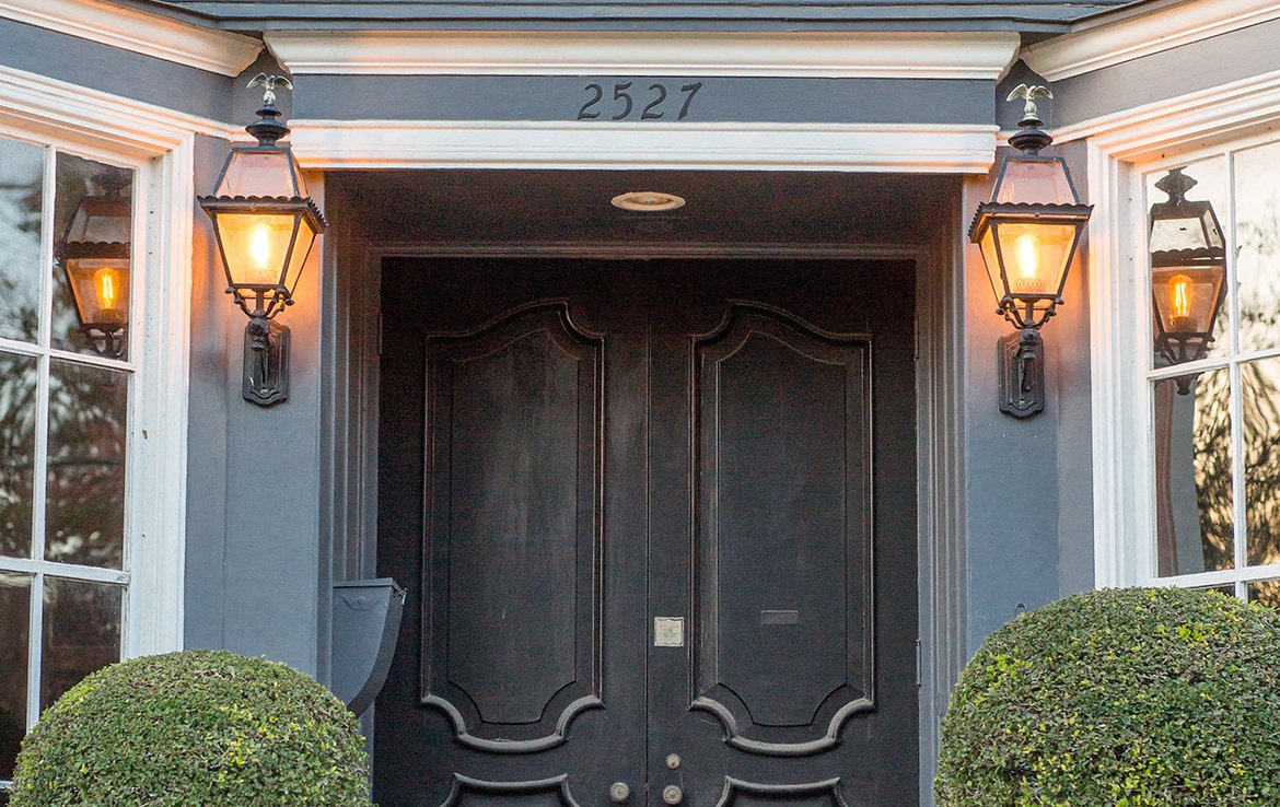 A double front door painted black with elegant molding and two lit wall lanterns on either side. The house number "2527" is displayed above the door. Two symmetrically trimmed bushes are in the lower front frame of the image, exemplifying a project from our portfolio at Orand Capital.