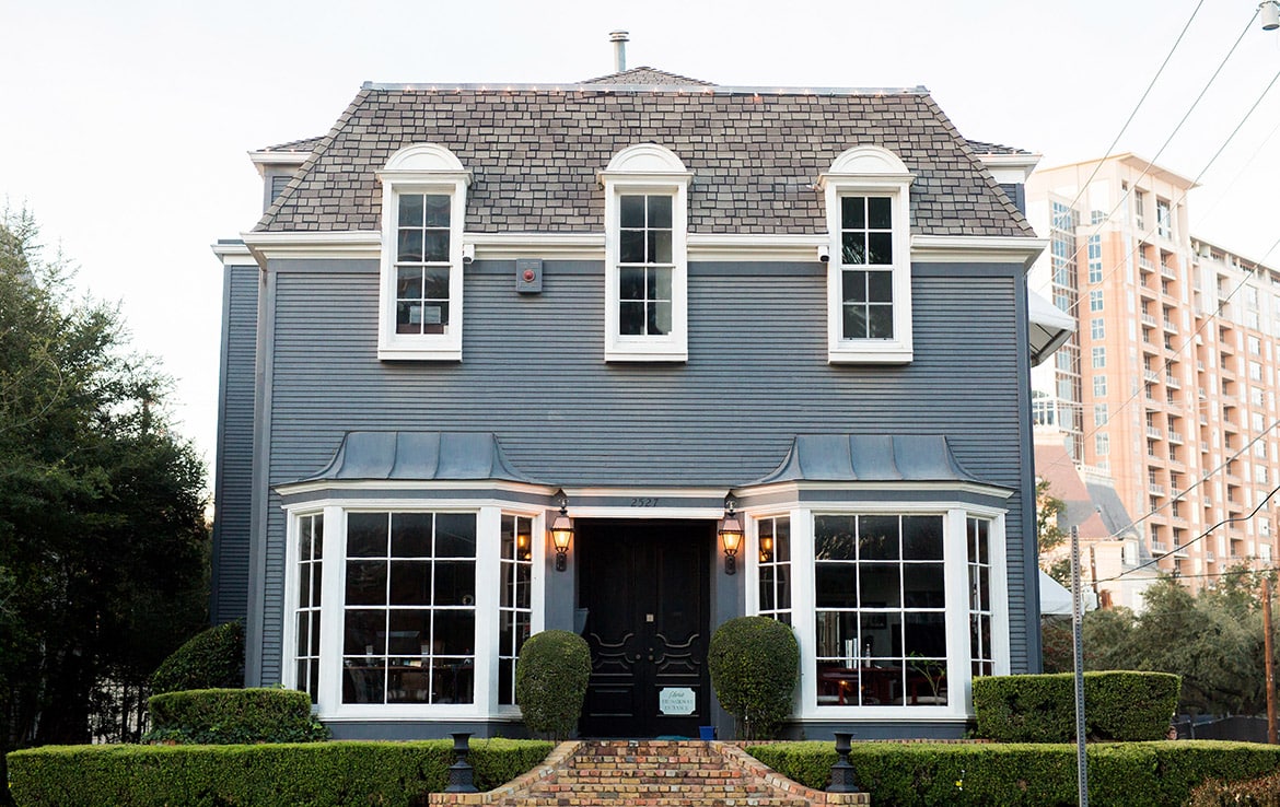 A two-story house with blue siding and a complex gabled roof graces our portfolio at Orand Capital. It features large front windows, a central black door with an ornate design, and is flanked by trimmed hedges. A brick pathway and steps lead to the entrance, with tall buildings visible in the background.