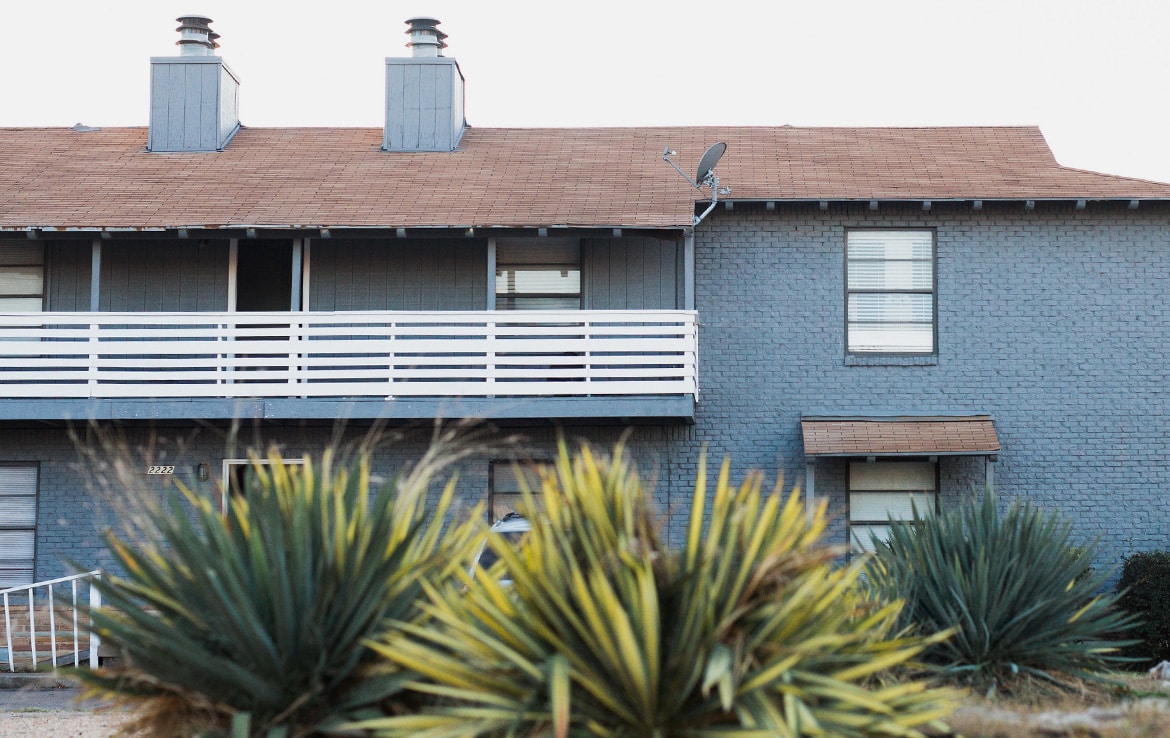 A two-story building with a grayish-blue facade and a brown roof. The upper level has a balcony with a white railing, and a satellite dish attached. In the foreground, there is a cluster of green and yellow spiky plants, perfectly showcasing another property in our portfolio - Orand Capital.