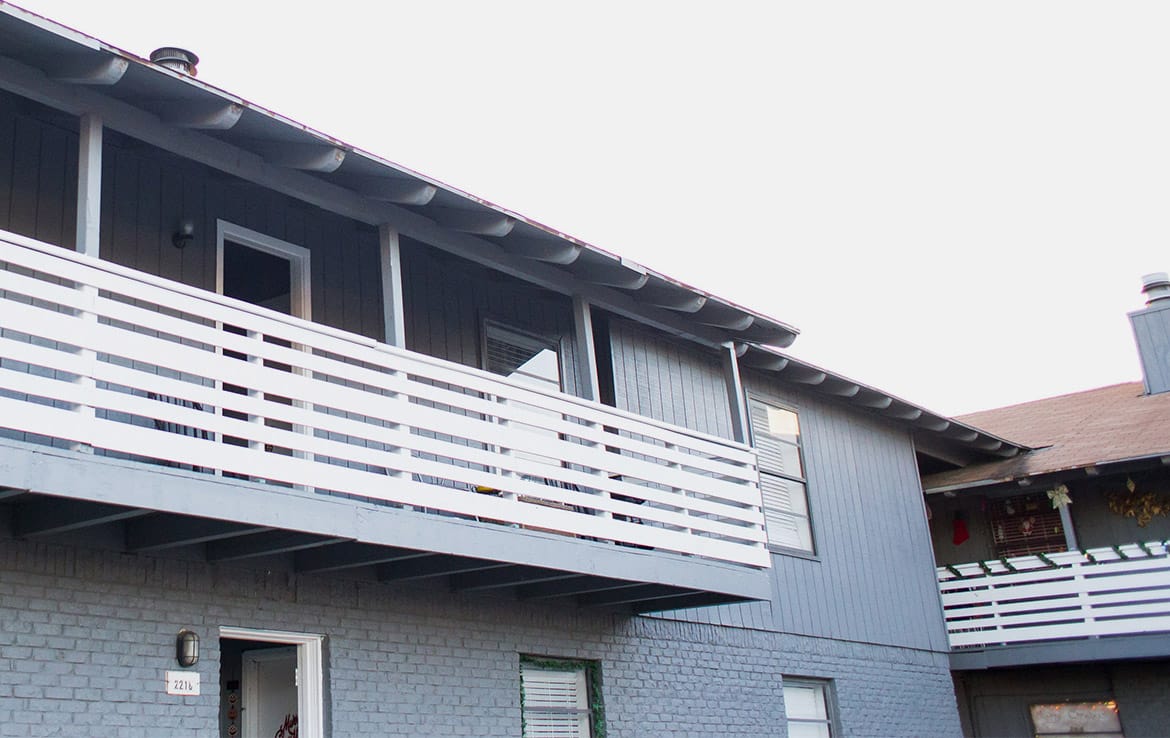 A two-story building with light gray siding and a white railing along the second-floor balcony. The building has a sloped roof and multiple windows. The entrance door on the ground level is open. Another similar building is partially visible in the background, illustrating Our Portfolio - Orand Capital.