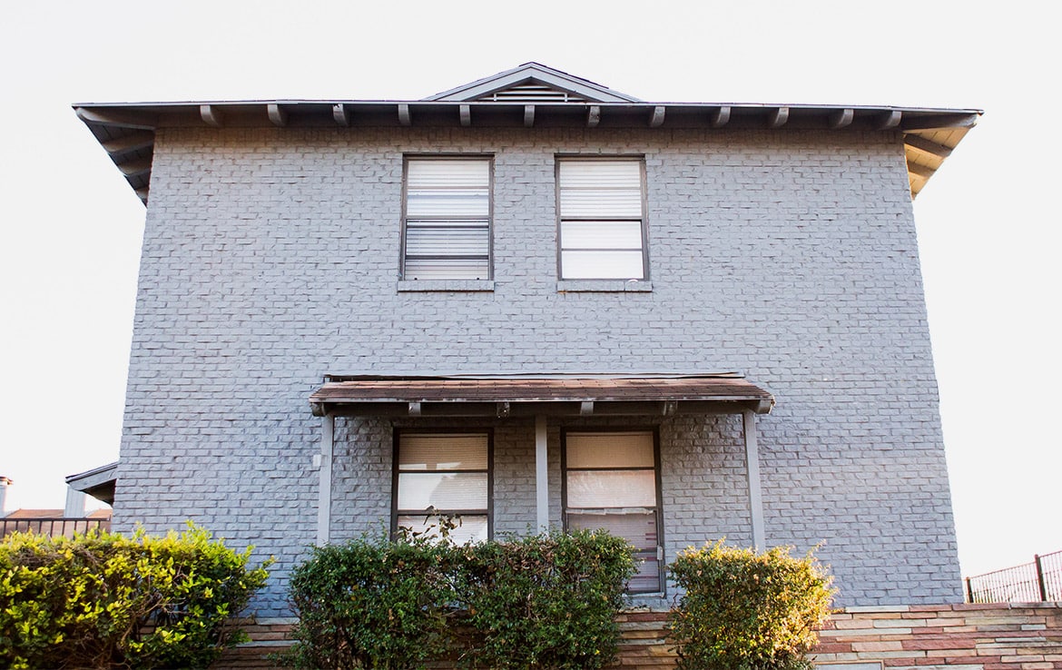 A two-story house from Our Portfolio features a light gray brick exterior, four front-facing windows, and a cozy porch with a wooden roof. Small bushes and a sloped stone pathway enhance the front yard's charm. The roofline gracefully extends beyond the walls, showcasing Orand Capital's meticulous design.