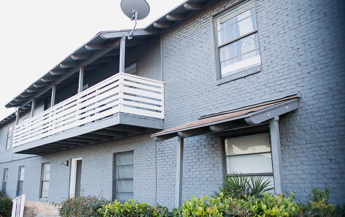 A two-story building with gray brickwork and a white balcony railing on the upper floor stands out in our portfolio at Orand Capital. There is a satellite dish mounted near the balcony. Trimmed shrubbery and plants are visible in front of the building, and the windows have white blinds.