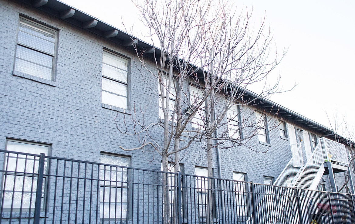 A two-story brick building with several windows stands behind a black metal fence and bare trees, showcasing an asset from Our Portfolio - Orand Capital. An exterior staircase on the right side leads to the second floor under a clear sky.