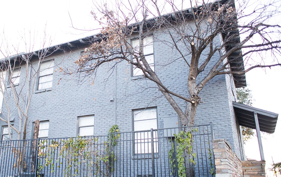 A two-story building painted gray with multiple windows is seen. The building has leafless trees in front and a black metal fence with greenery growing on it. A partially visible staircase on the right side leads to an entrance, showcasing a prime example from our portfolio at Orand Capital.