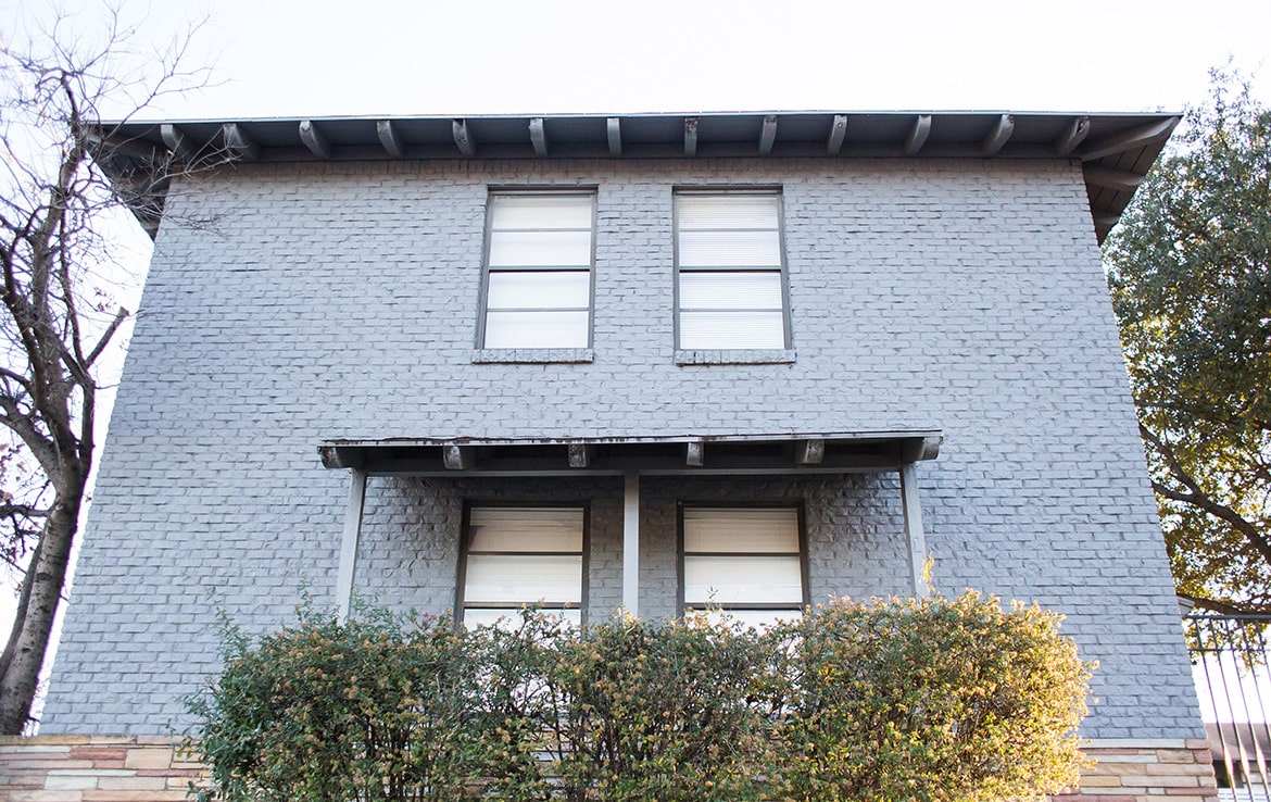 A two-story gray brick building with two windows on each floor. The windows are covered with white shades. A roof overhang protects the entrance, which is flanked by bushes. The sky is clear and trees are visible on the sides of the building, beautifully presented in our portfolio at Orand Capital.