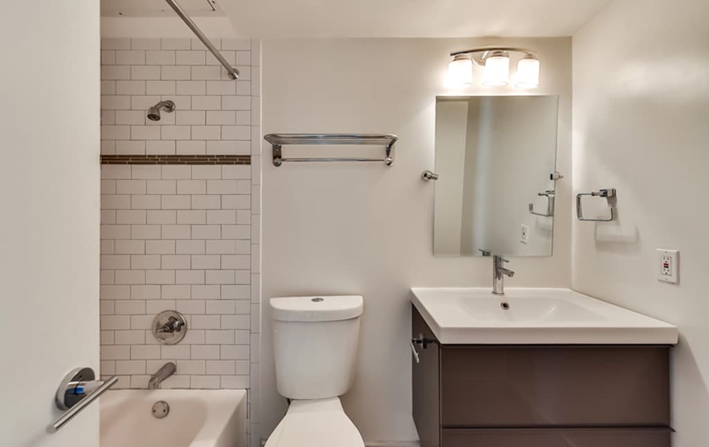 A modern bathroom from our portfolio featuring a white bathtub with a showerhead, white subway tile walls, and a sleek vanity with a large rectangular mirror and three-light fixture above. The room also includes a white toilet, towel rack, and single-handle faucet on the sink — Orand Capital design.