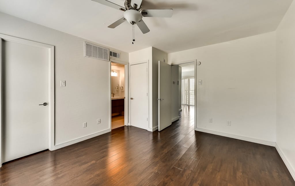 A bright, empty room with wooden floors, light-colored walls, and a ceiling fan. There are two doors; one leads to a bathroom with visible sink and mirror, and another door is open, leading to an outside area through vertical blinds. This space is featured in our portfolio at Orand Capital.