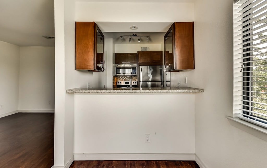A modern kitchen with stainless steel appliances, dark wood cabinets, and a granite countertop. The kitchen is seen through a pass-through window with a counter ledge. The room has dark hardwood floors and is adjacent to a living space and a bright window with blinds. Featured in Our Portfolio - Orand Capital.